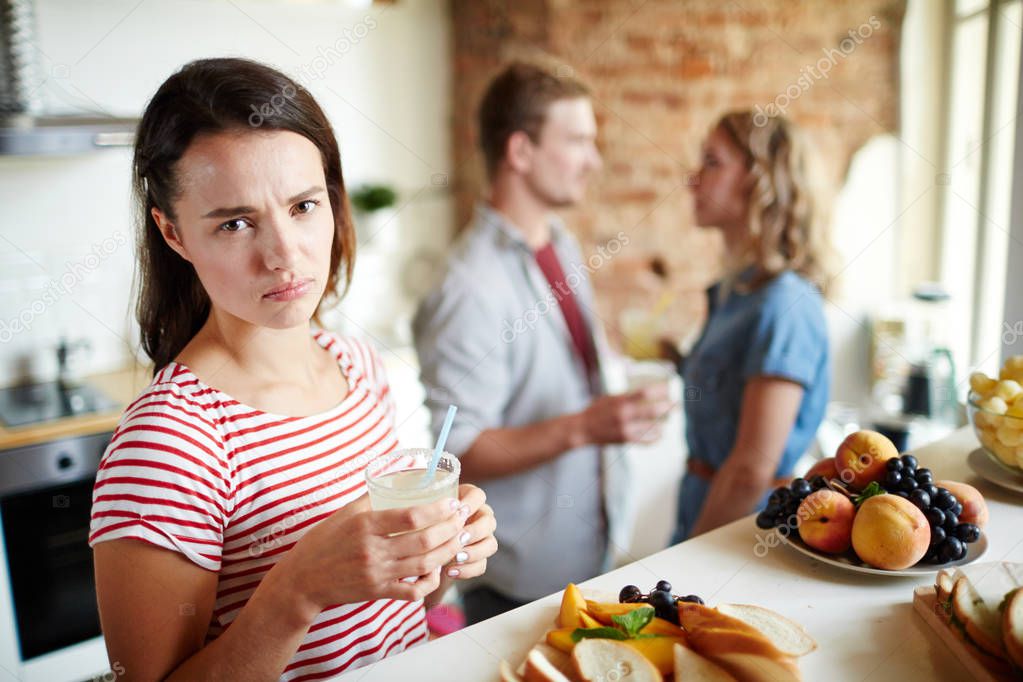 Jealous girl with homemade drink looking at camera on background of young couple having conversation