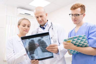 İki doktor akciğer röntgenini tartışırken genç stajyer kliniklerde durup notlar alıyor.