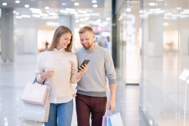 Akıllı telefonlu neşeli kız ve erkek arkadaşı alışveriş merkezinde takılırken internet ürünlerini karıştırıyor.