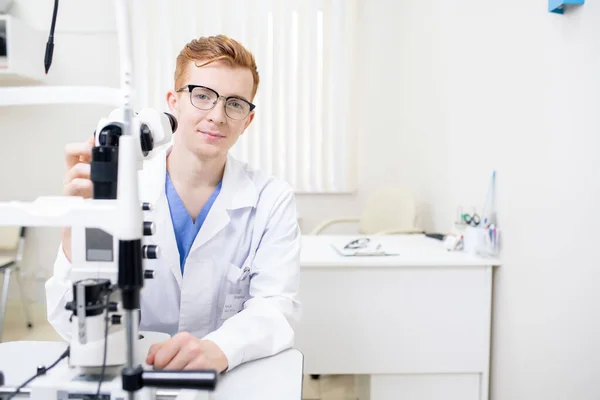 年轻的成功眼科医生穿着制服坐在工作场所 同时在工作场所测试新的眼科设备 — 图库照片