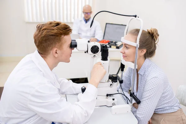 女性患者の前で視力検査装置のレンズを通して見る白衣姿の若い検眼士 — ストック写真