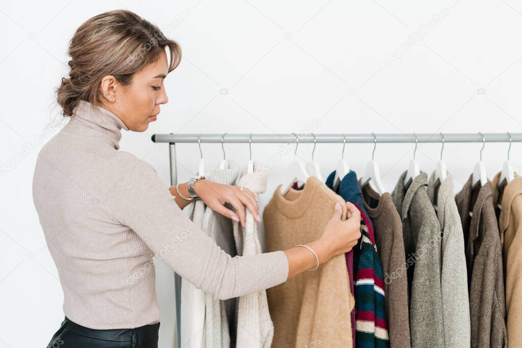 Casual young woman looking at beige sweater on rack among other clothes while going to buy something