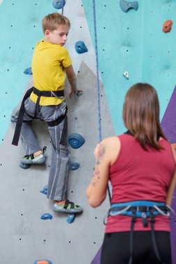 Sarı tişörtlü ve gri pantolonlu aktif bir çocuk eğitmen tavsiyesini dinlerken eğitim sırasında duvara küçük kayalarla tırmanıyor.