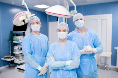Team of three contemporary surgeons in masks, gloves and protective uniform standing in front of camera in hospital clipart