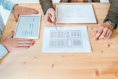 Kağıt ve dokunmatik ekranda yeni web sitelerinin planlarını tartışan iki genç tasarımcının elleri