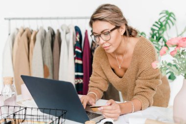 Günlük bej rengi kazaklı genç bir kadın çevrimiçi dükkanda sipariş verirken dizüstü bilgisayarına bakıyor.