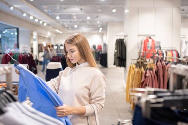 Güzel genç bayan alışverişçi yeni gömleklerle askıda duruyor ve günlük kıyafet reyonunda mavi olana bakıyor.