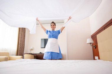 Üniformalı ve önlüklü genç oda hizmetçisi sabah işini yaparken beyaz temiz çarşafları yatağın üstünde tutuyor.