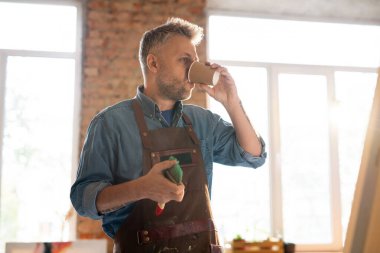 İş kıyafetleri içinde orta yaşlı bir ressam, teneffüste sıcak kahve içerken iş yerinin yanında boyama atölyesinde duruyor.