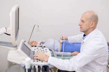 Ciddi doktor ultrasonografi cihazlarıyla hastanın karın bölgesini incelerken ekrana bakıyor.