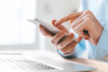 İş yerinde mobil cihazlar kullanırken dizüstü bilgisayarın üzerindeki akıllı telefon ekranını gösteren genç işadamının elleri
