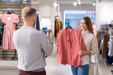 Akıllı telefonlu genç adam, kız arkadaşının elinde% 50 indirimli pembe ceketiyle alışveriş merkezinde fotoğraf çekiyor.