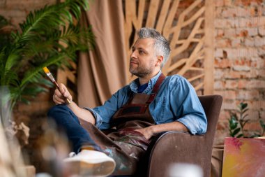 Kır saçlı, boyalı fırçalı dalgın bir ressam koltukta oturuyor ve stüdyoda resim yaparak sehpaya bakıyor.