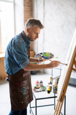 İş kıyafetlerinde çalışan çağdaş ressam palet ve boya fırçasıyla resim yaparken sehpanın önünde duruyor.