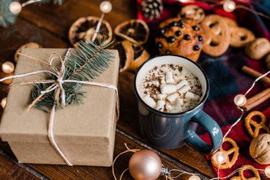 Marşmelovlu sıcak kahve bardağı, kozalaklı hediye kutusu, ahşap masada kurabiye ve çelenk.