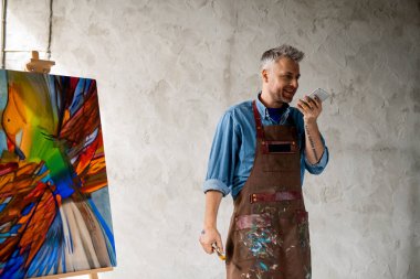 Çalışan neşeli orta yaşlı ressam stüdyoda duvarın önünde dikilirken akıllı telefondan sesli mesaj kaydediyor.