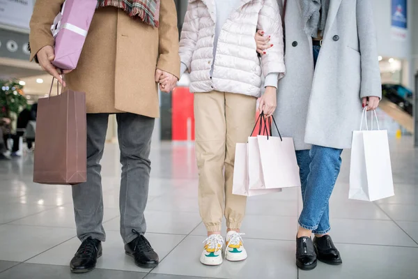 Lower part of casual family of three holding paperbags with purchases while spending time in the mall after shopping