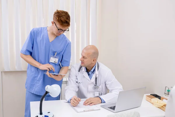 Junge Ärztin Blauer Uniform Präsentiert Reiferen Arzt Beim Scrollen Touchpad — Stockfoto