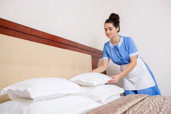 かなり若いですブルネット女性で制服を着てホテルの部屋でベッドを作りながら白い新鮮な枕 — ストック写真