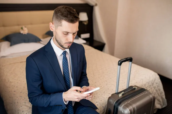 ホテルの部屋でベッドに座っている間にタクシーを呼び出すためにスマートフォンの連絡先を介して見てエレガントなスーツの若い重大なビジネスマン — ストック写真