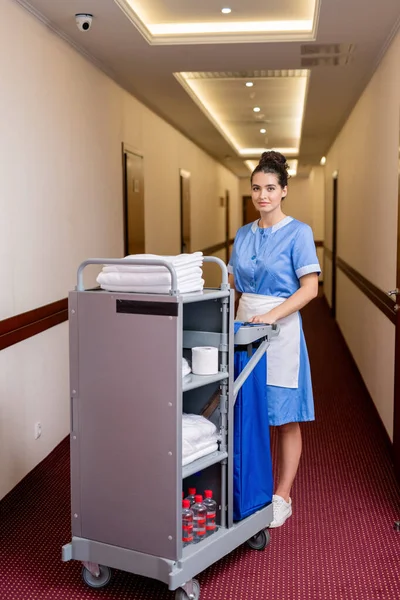 きれいなタオルやホテルの部屋で他のものを取得しながら 長い廊下に立って制服を着たかなり若い部屋のメイド — ストック写真