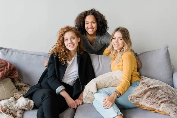 Group of friendly intercultural girls in smart casual sitting on sofa in front of camera while enjoying home rest