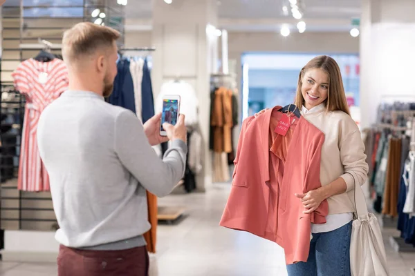 Happy girl with toothy smile holding pink jacket by chest while standing in front of young man photographing her on smartphone