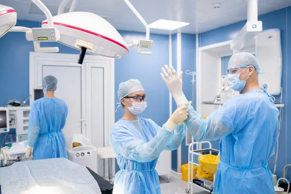 年轻的助手在医院准备动手术时把橡胶手套戴在外科医生的手上 — 图库照片