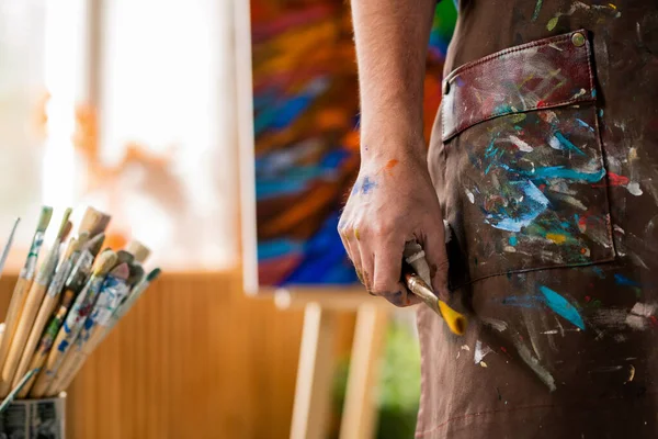 自身のアトリエで制作しながら 絵筆を持つエプロンの現代美術家の手 — ストック写真