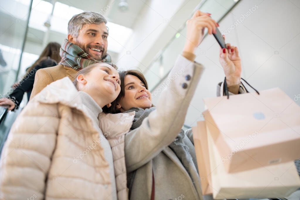 Happy young contemporary family of three making selfie after shopping while moving down on escalator in the mall
