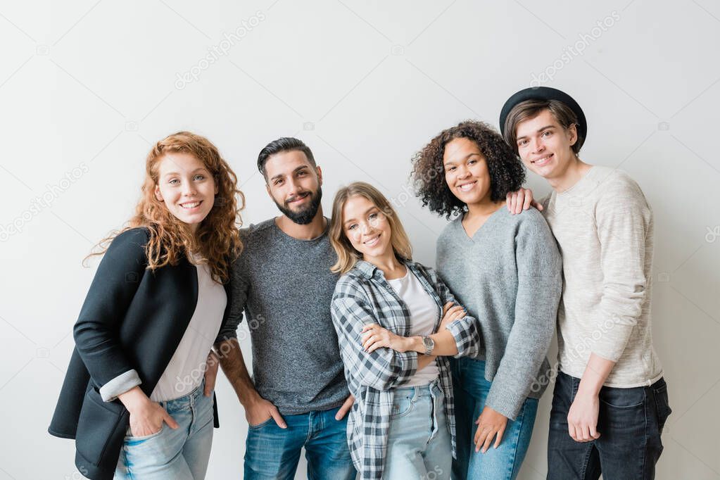 Friendly smiling guys and girls in casualwear standing by white wall in front of camera and looking at you