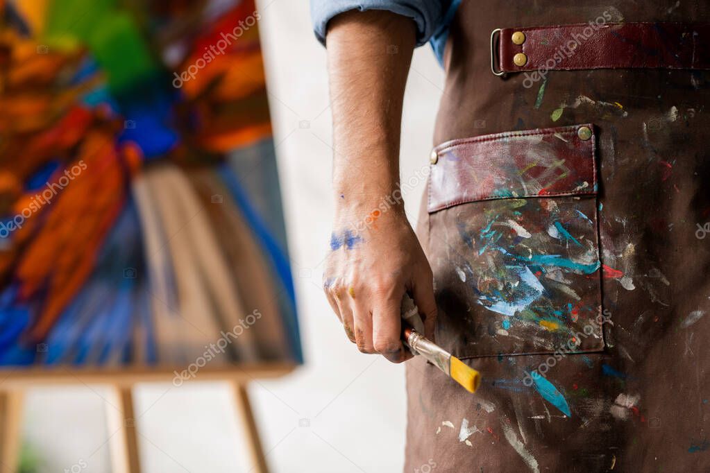 Part of professional painter in dirty apron holding paintbrush in hand while standing on background of painting on easel