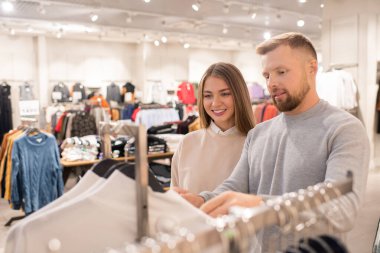 Neşeli kız ve erkek arkadaşı alışveriş merkezindeki ya da alışveriş merkezindeki günlük kıyafet reyonundaki kazaklara ya da kazaklara bakıyor.