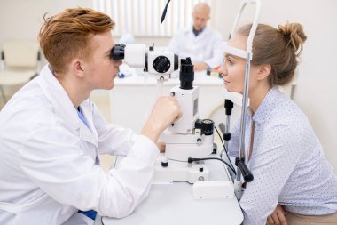 Göz doktoru ve hastası optometrik ekipmanları kontrol ederken birbirlerinin önünde oturuyorlar.
