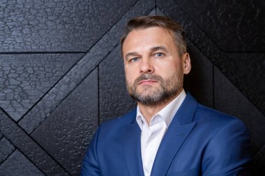 Ciddi, olgun, orta yaşlı, sakallı bir iş adamı, siyah ahşap duvarın önünde duruyor.