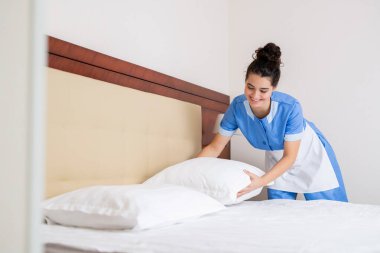 Oda hizmetçisi üniformalı mutlu esmer kız otel odasında işini yaparken beyaz temiz yastıkları yatağa koyuyor.