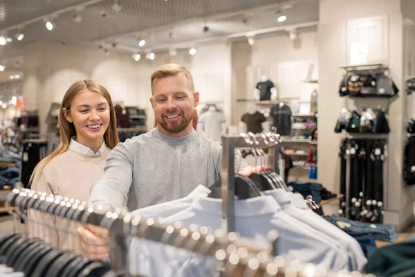 Genç ve mutlu bir çift, erkek için yeni bir tane seçerken, günlük kıyafet reyonunda gömleklerle askıda bekliyor.