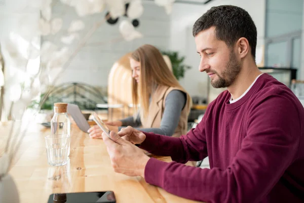 現代の若いモバイル男は 女性を背景にカフェでテーブルで休んでいる間にデジタルタブレットでスクロールします — ストック写真