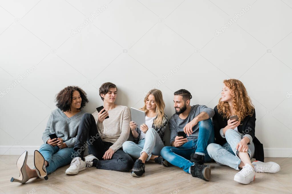 Happy young intercultural friends with gadgets sitting in row by wall and watching funny video in touchpad held by blonde girl