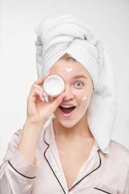 Kafasında havluyla, kameranın önünde bir gözünün yanında yüz kremi kavanozu tutan güzel, kendinden geçmiş genç bir kadın.