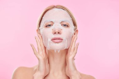 Genç bir kadın kameranın önünde cildini korurken yüzüne ferahlatıcı tekstil maskesi takıyor.