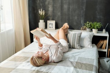 Hafta sonları evde otururken pijamalarıyla yatakta dinlenen ve kitap okuyan güzel genç bir kadın.