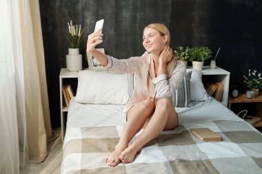 Pijamayla gülümseyen genç bir kadın sabahları evde pencerenin önünde yatarken selfie çekiyor.