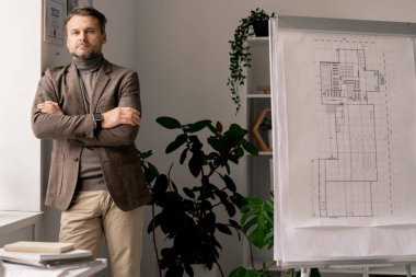 Ciddi bir mimar ya da mühendis kollarını göğsünde kavuşturup ofis penceresi ile beyaz tahtanın arasında duruyor.