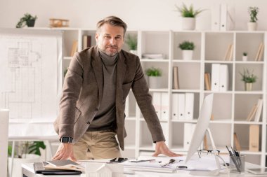 Successful and confident architect in formalwear bending over desk with computer monitor and office supplies clipart