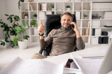 Akıllı telefonlu neşeli bir mimar ya da iş adamı ofiste otururken bir müşteriyle ya da meslektaşıyla konuşuyor.