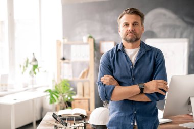 Sakallı mühendis, iş yerinde kameranın önünde dikilirken göğsünde çapraz kollar var.