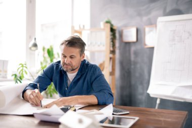 Günlük giyimde olgun bir mimar masa başında otururken yeni çizim sırasında kalemle plan üzerinde çizim yapıyor.