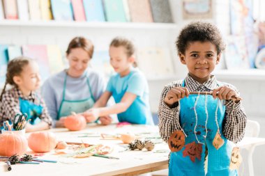 Mavi önlüklü Afrikalı okul çocuğu elinde iplikte asılı kuru yapraklardan yapılmış cadılar bayramı süslemeleriyle sopa tutuyor.
