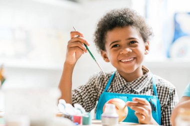 Boyalı fırçalı neşeli okul çocuğu derste Noel oyuncağı topunu yaparken hediyeyi kime vereceğini düşünüyor.